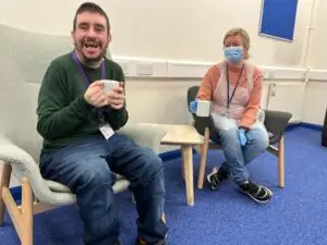 A person supported by Sense Scotland sits on a chair holding a mug, and smiles at the camera next to his support worker