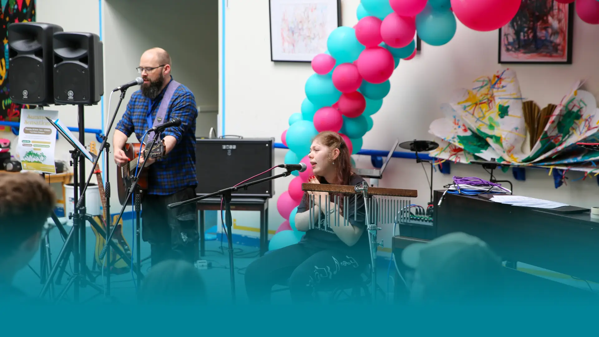 A man in a blue shirt plays guitar while a girl sits on a chair next to him and sings in front of a balloon arch