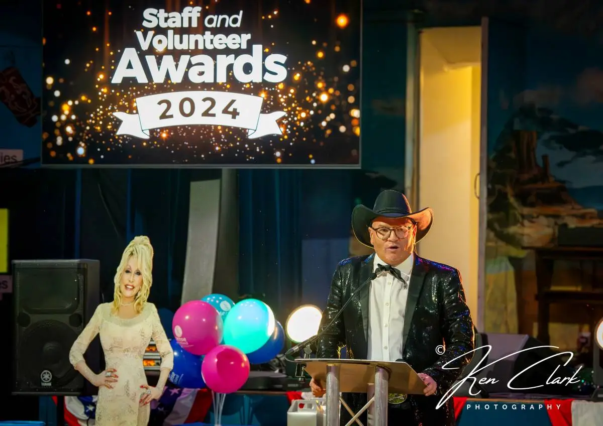 a man in a glittery suit and a cowboy hat stands at a lectern next to a cardboard cutout of dolly parton