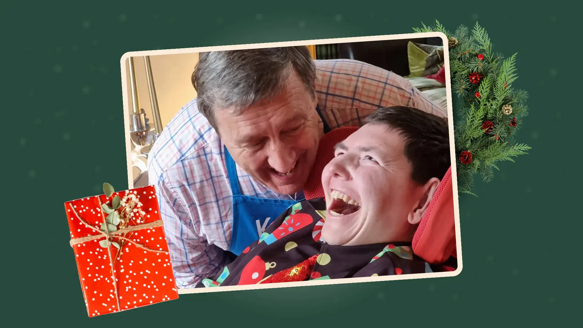 A young man smiling wearing a Christmas jumper and his father, also smiling, looking at him