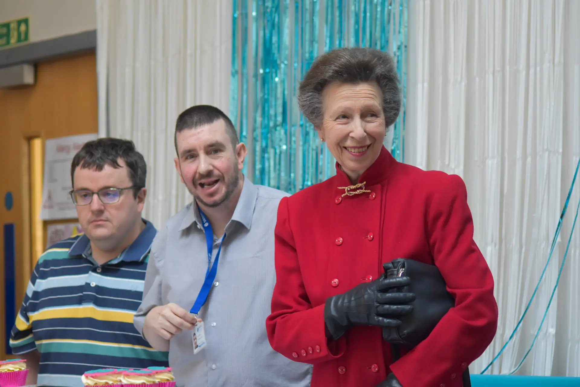 The Princess Royal in a red coat with black gloves, smiling to the right of the camera. To her left are two people supported by Sense Scotland