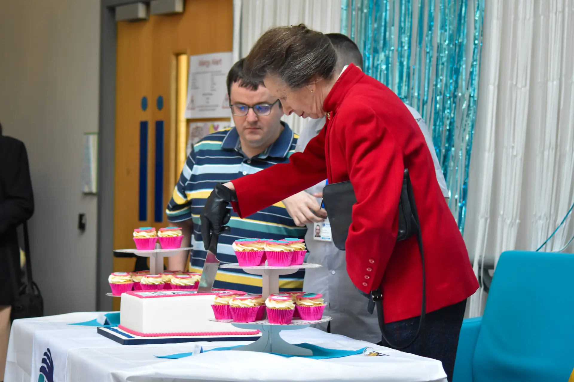 Princess Anne cuts a white cake with pink trims. Next to the cake are two people supported by Sense Scotland and cupcakes with pink cases.