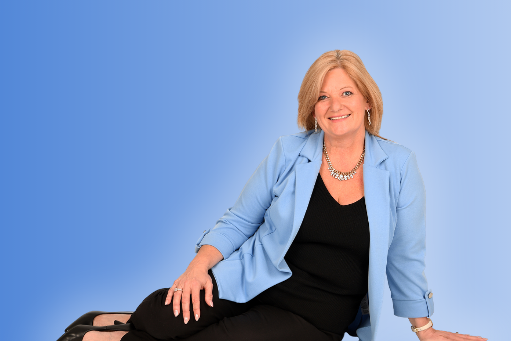 A blonde woman in a light blue blazer smiles at the camera while sitting on the floor in front of a blue background.