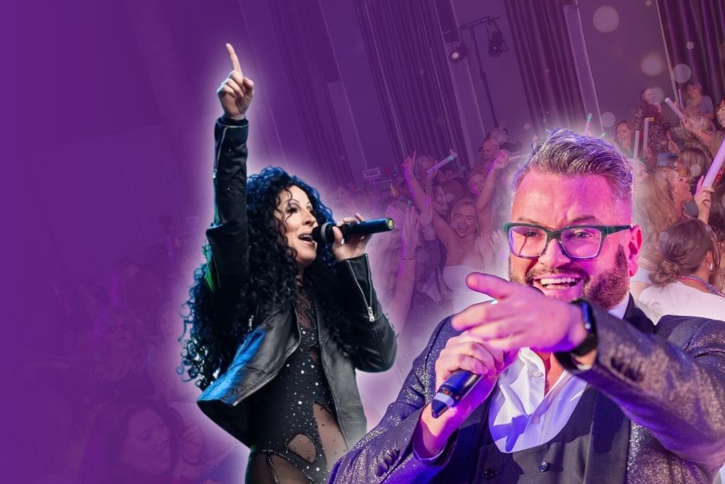 A man wearing a suit and glasses speaks into a microphone and points into the crowd. Next to him, a Cher tribute act dressed in black sings into a microphone and holds her hand up to the sky.