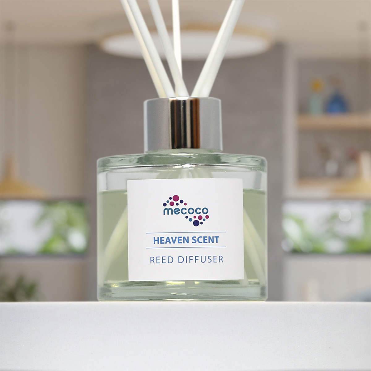 A glass reed diffuser with a sticker featuring the Mecoco logo and text which says 'Heaven Scent Reed Diffuser'. Behind it you can see a blurred living space.