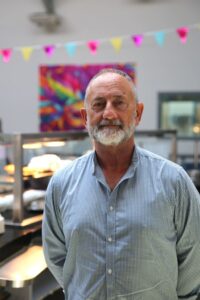 A man in a check shirt stands in a large room. Behind him are colourful flags and heating lights for a kitchen.