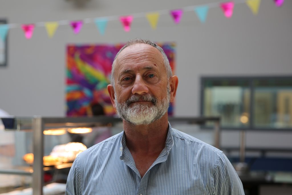 A man in a check shirt stands in a large room. Behind him are colourful flags and heating lights for a kitchen.