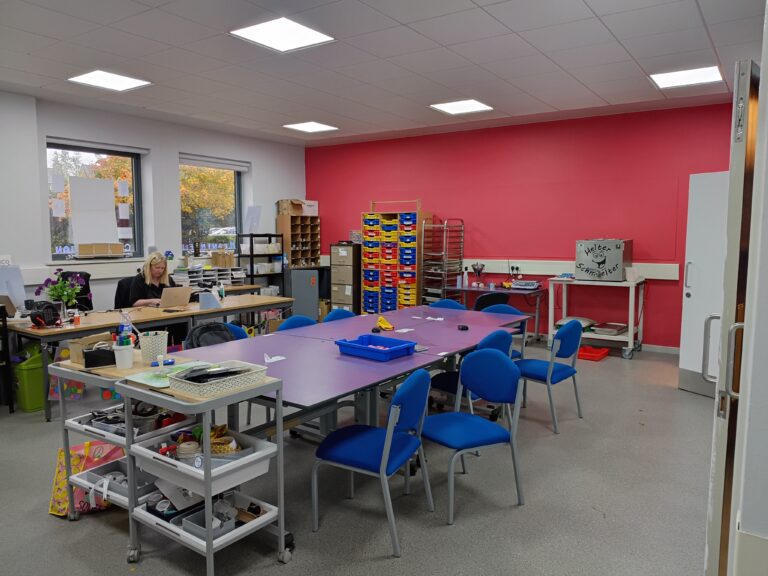 A workshop space with a large purple table at the centre of it and chairs around it. One wall at the far end is painted red. The wall on the left has two windows, and a woman is sat at a desk in front of them.